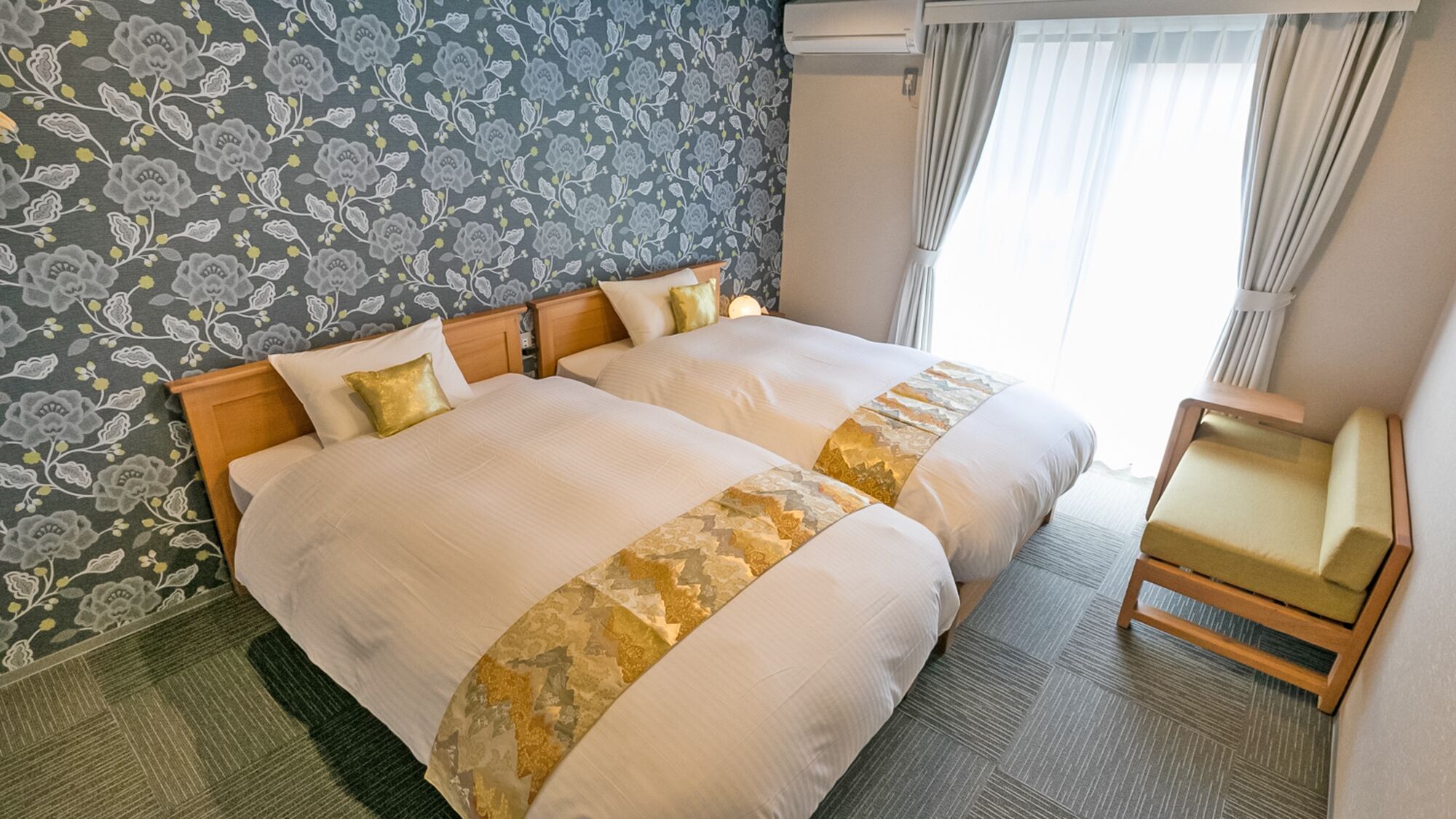 Uma cama de casal com cabeceira estampada em flor, uma mesinha de cabeceira e uma cadeira.