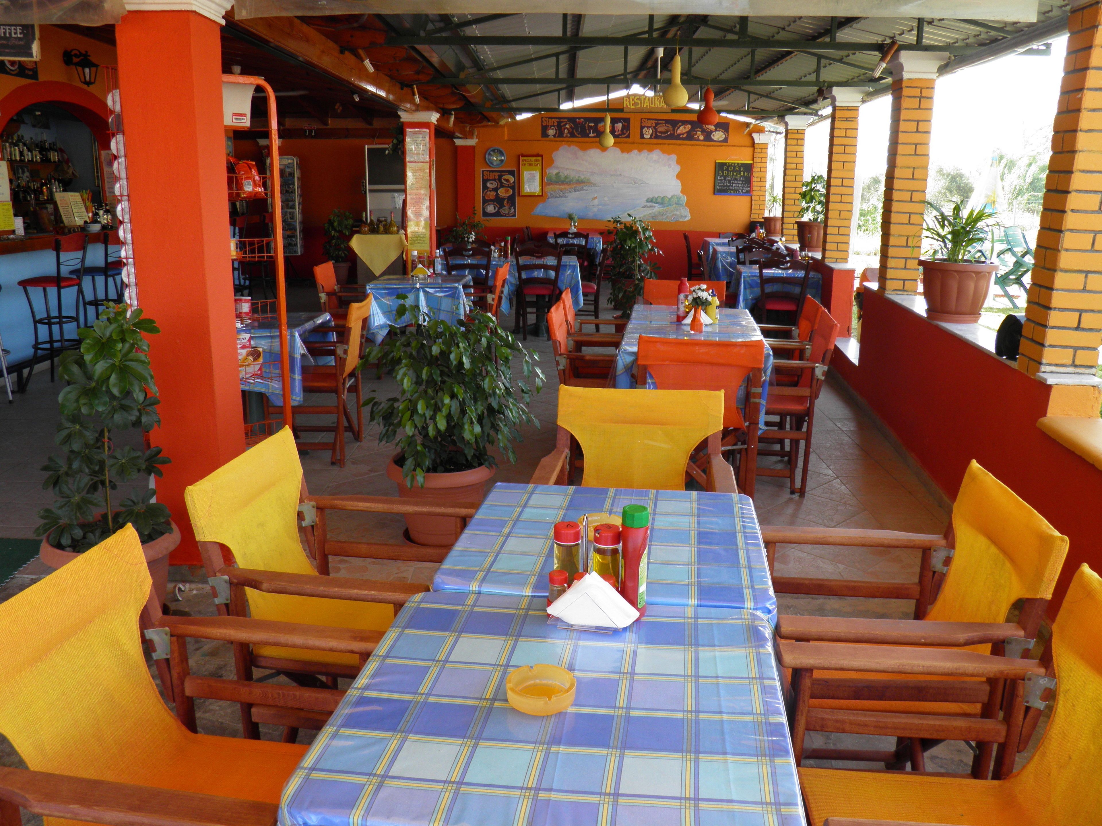 An outdoor restaurant with tables covered in blue and yellow checkered tablecloths, surrounded by orange chairs and potted plants.