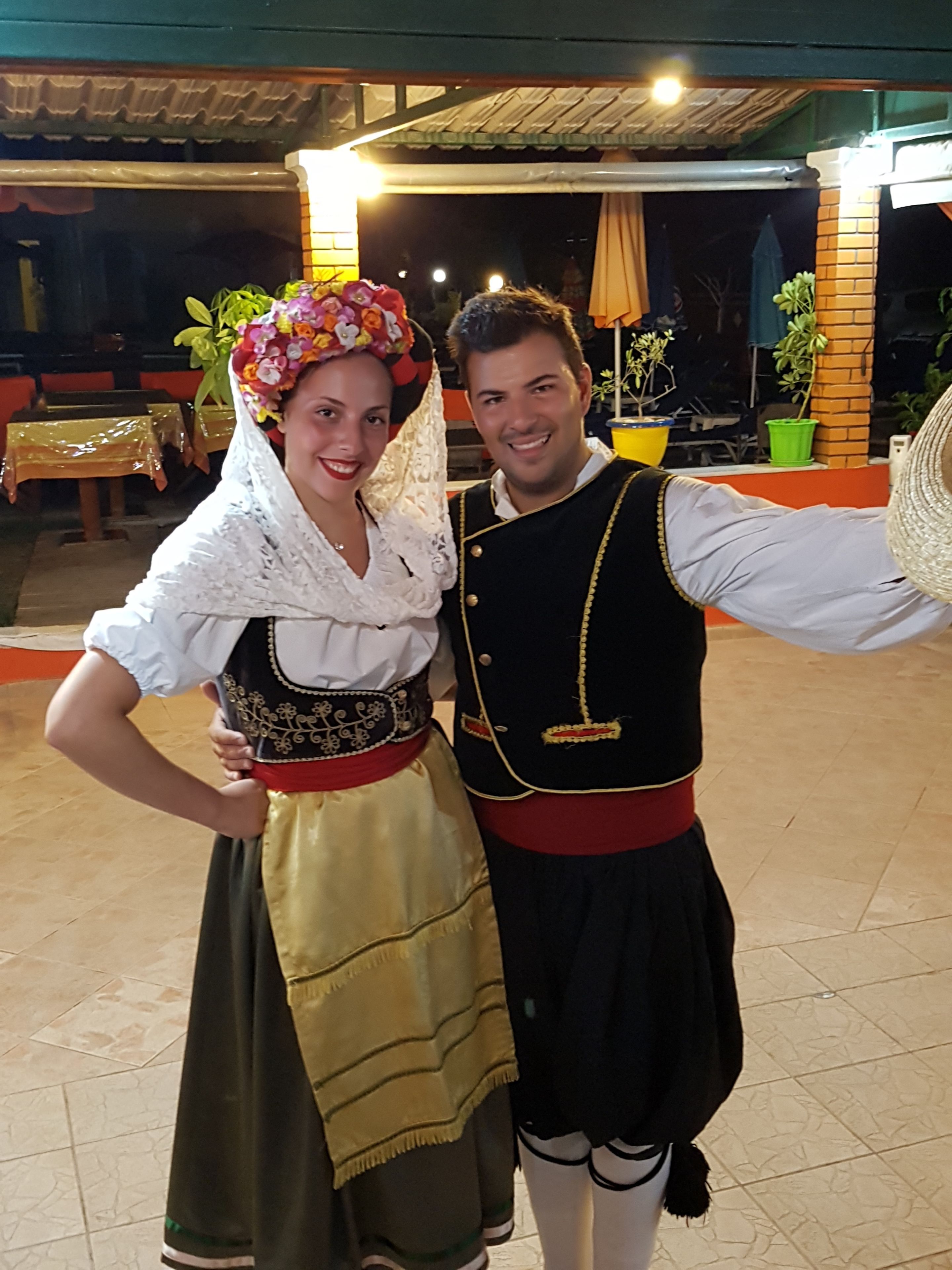 A couple dressed in traditional attire posing in an outdoor setting with a tiled floor and a brick wall in the background.