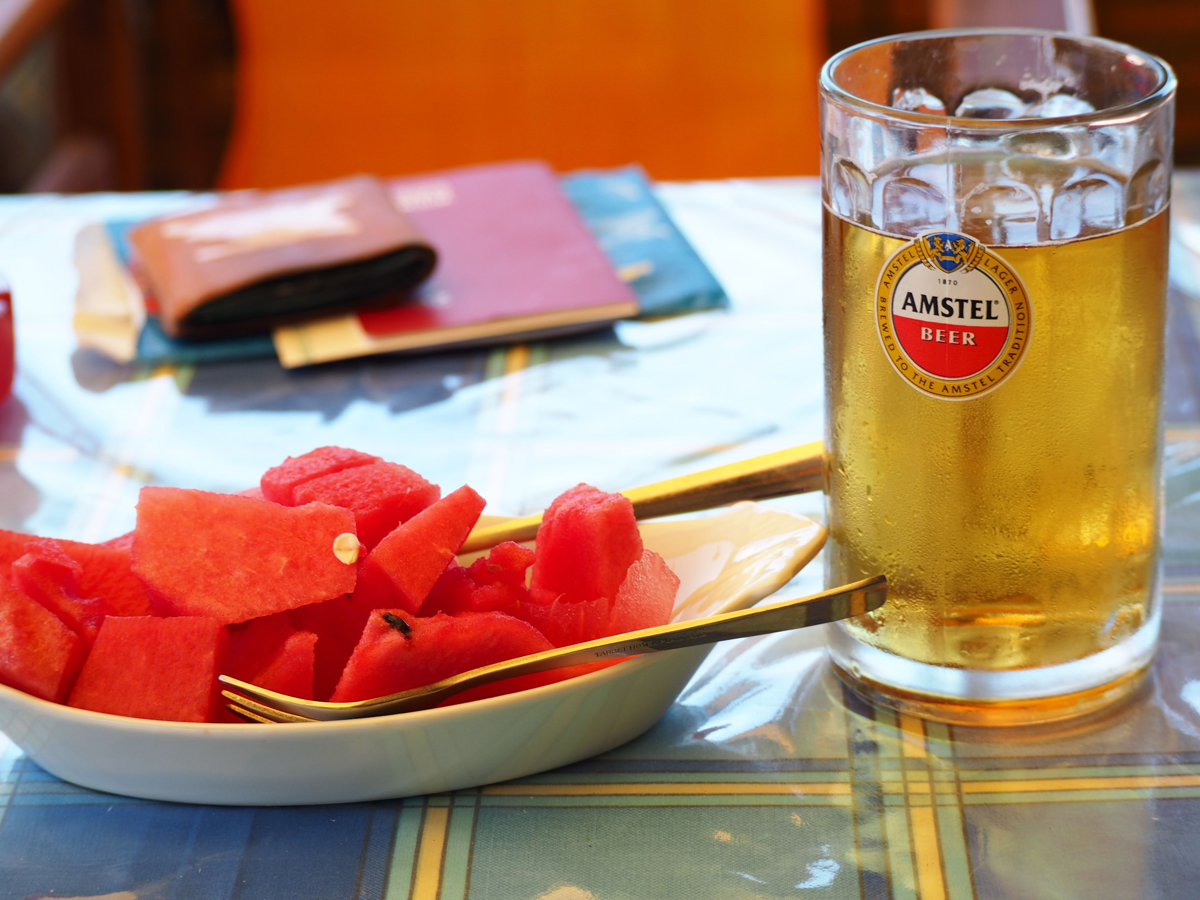 Una cerveza en vaso con etiqueta, un tazón de cubos de sandía y una cuchara sobre la mesa.