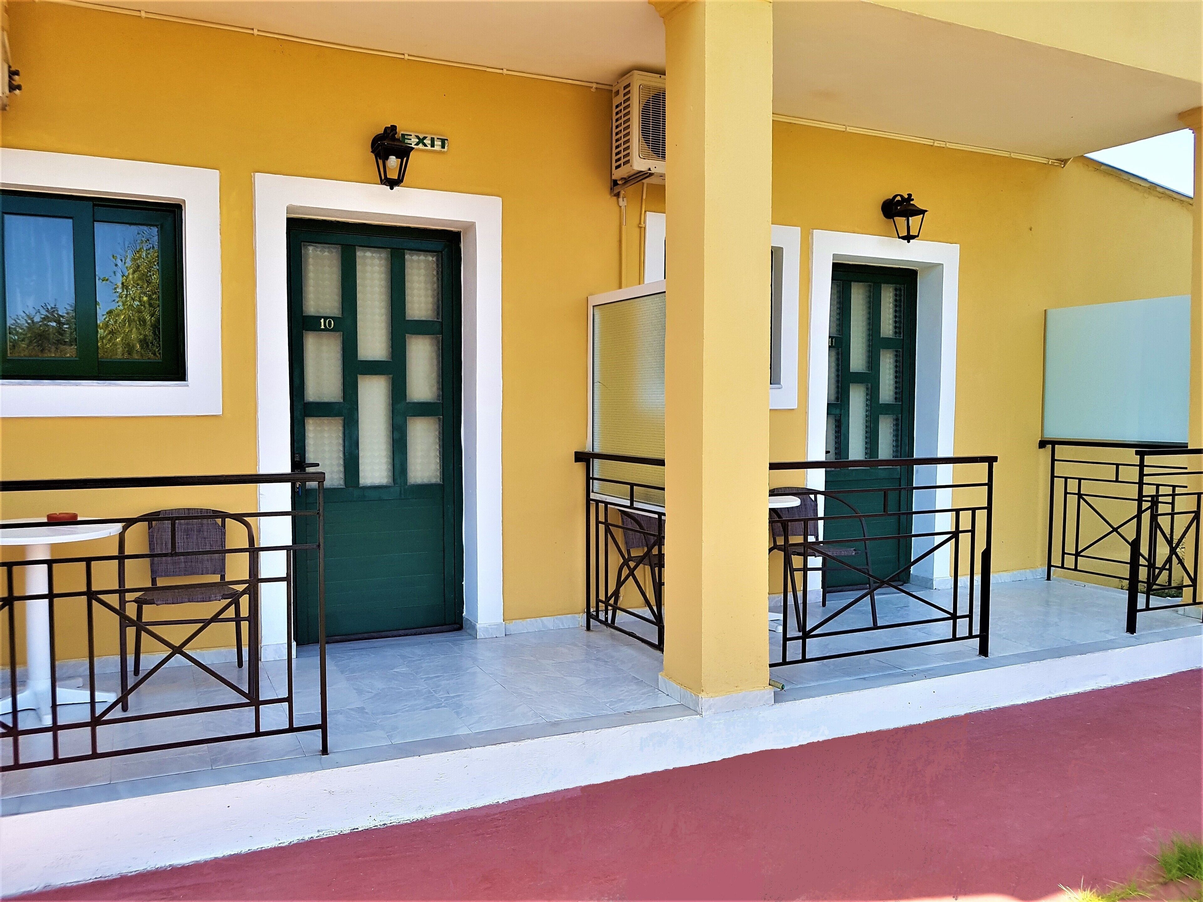A yellow building with a green door, a white door, and a window. There are two outdoor tables with chairs.