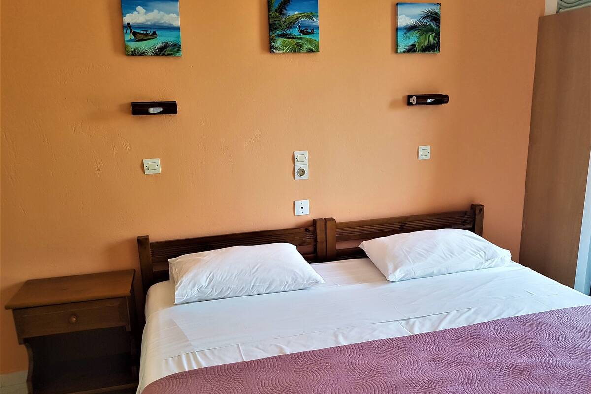 A bedroom with a wooden bed, white pillows, a purple bedspread, and three wall-mounted paintings of tropical scenes.