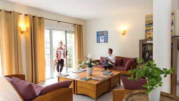 Une pièce avec un canapé en bois, une table basse avec des magazines, une plante en pot et une porte-fenêtre coulissante donnant sur l’extérieur.