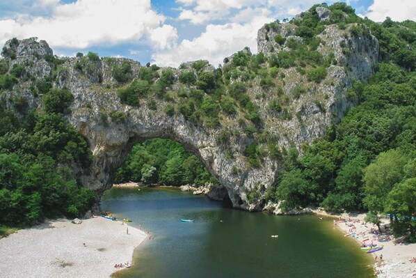 A natural rock arch formation over a body of water with a sandy beach and greenery on the sides.