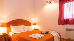 Une chambre d’hôtel avec une tête de lit en bois, deux lampes de chevet, un lit avec du linge de lit blanc, un plateau avec un verre d’eau et un croissant, et une fenêtre avec des rideaux rouges.