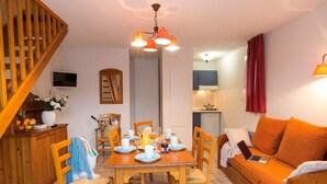 Un salon chaleureux avec un escalier en bois, un coin repas avec une table dressée pour quatre personnes, et une cuisine en arrière-plan.