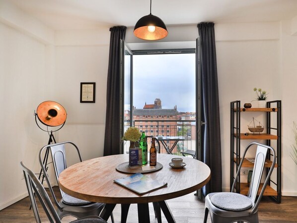 A round dining table with chairs, a hanging light fixture, a view of buildings through large windows, and a shelf with decorative items.