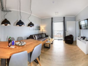 A modern living room with a sofa, coffee table, and a dining area with a wooden table and chairs. There is a TV on a stand and a balcony visible through the window.