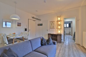 A modern living room with a grey sofa, a dining table with chairs, and a kitchen area visible through an open door.