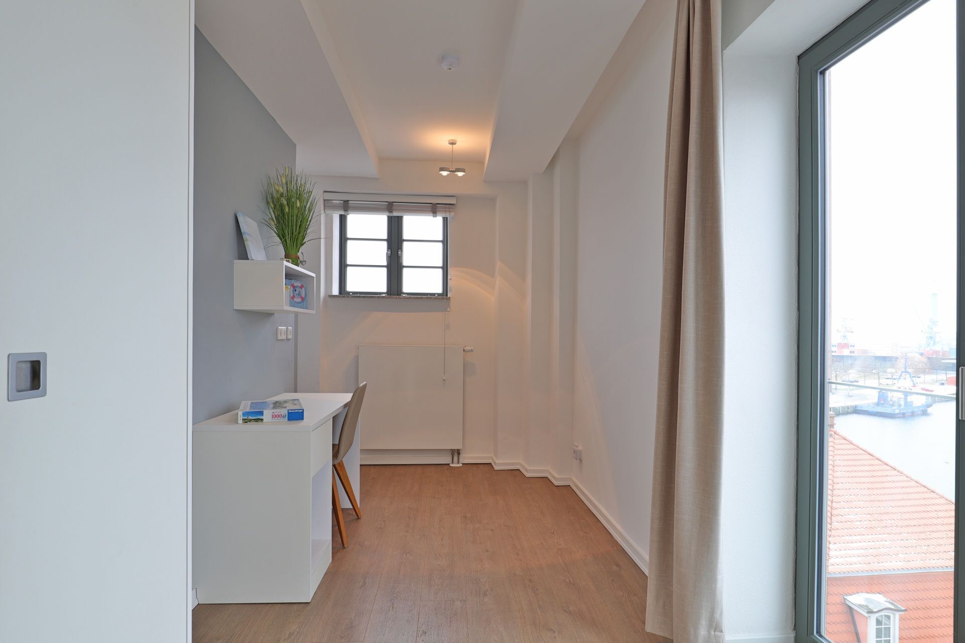 Een moderne, minimalistisch ingerichte kamer met uitzicht op een stadsgezicht door een groot raam, een radiator, een klein tafeltje met een stoel, en een plankje met decoratieve objecten.