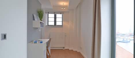 Ein modernes, minimalistisches Zimmer mit Blick auf eine Stadtansicht durch ein großes Fenster, inklusive Heizkörper, einem kleinen Tisch mit Stuhl und einem Regal mit Dekoration.