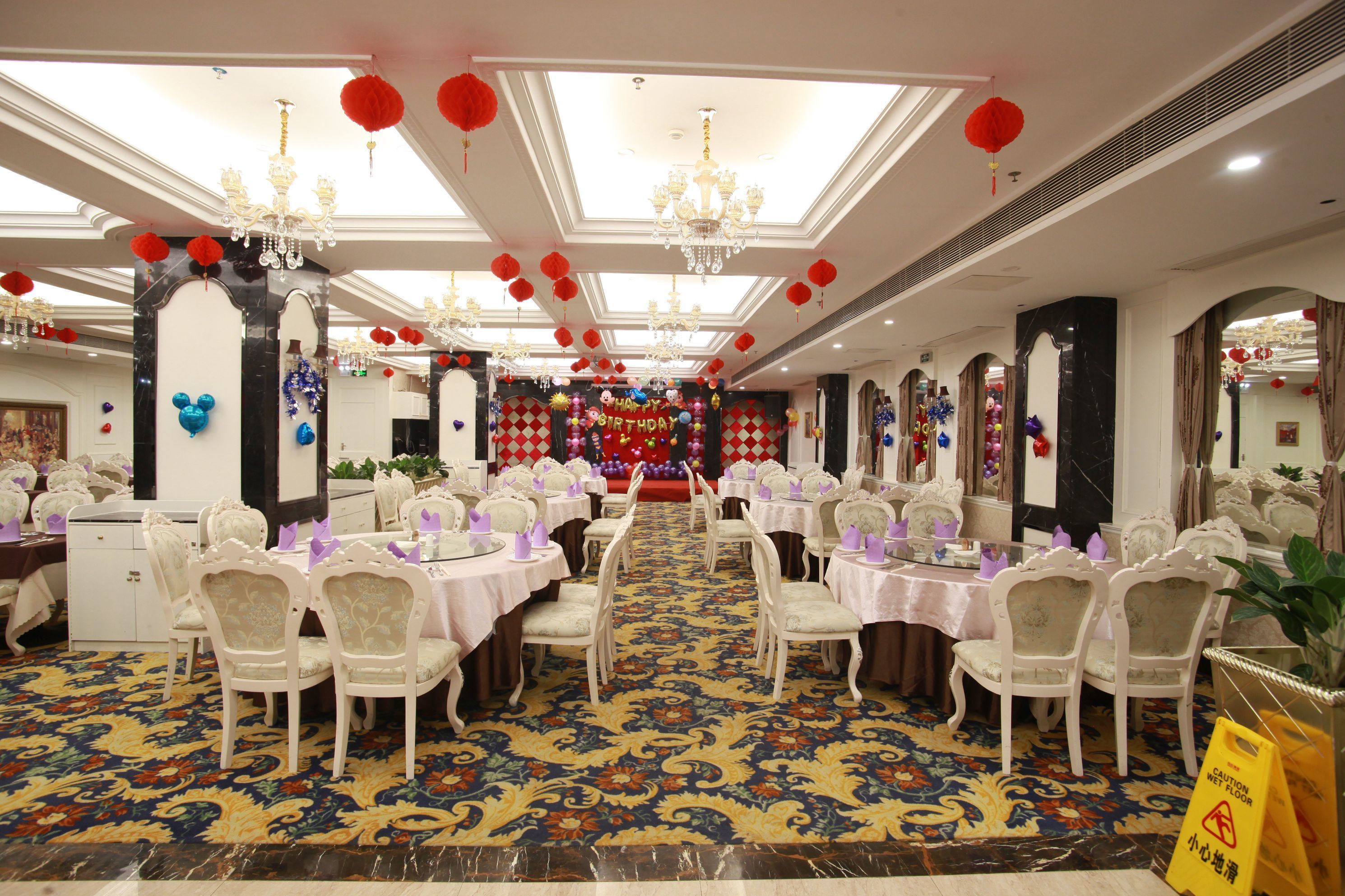 A banquet hall with round tables, white chairs, chandeliers, and red lanterns.
