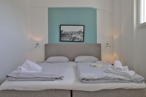 Ein Doppelbett mit zwei Kissen, eine türkis gestrichene Wand, ein gerahmtes Stadtbild und zwei Nachttischlampen.