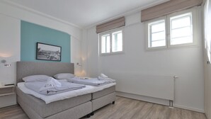 Une chambre à coucher comprenant un lit, une table de chevet, une lampe fixée au mur, une fenêtre avec des stores et un radiateur.