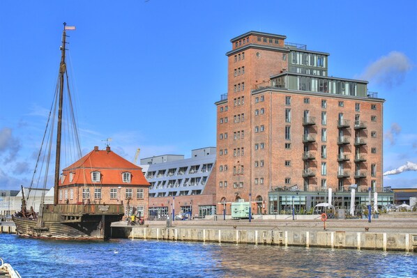 A waterfront building with a red-roofed structure, a tall brick building with balconies, and a dock with a boat. - Ohlerich storage app. 03 - Ohlerich Speicher App. 03 (Wismar)