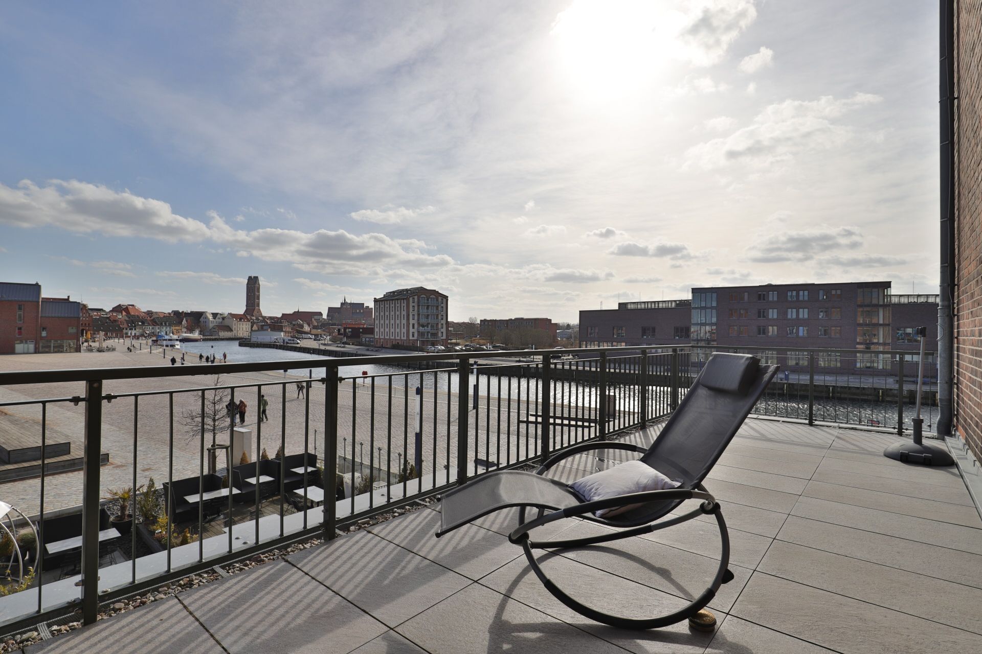 A balcony with a metal railing, a single lounge chair, and a view of a riverside cityscape.