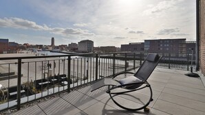 Un balcon avec une balustrade en métal, un fauteuil et une vue sur le paysage urbain qui s’étend le long de la rivière.