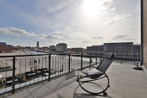 A balcony with a metal railing, a single lounge chair, and a view of a riverside cityscape.