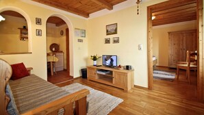 Ein gemütliches Wohnzimmer mit Holzmöbeln, einem Fernseher und einem durch einen Bogen sichtbaren Küchenbereich.