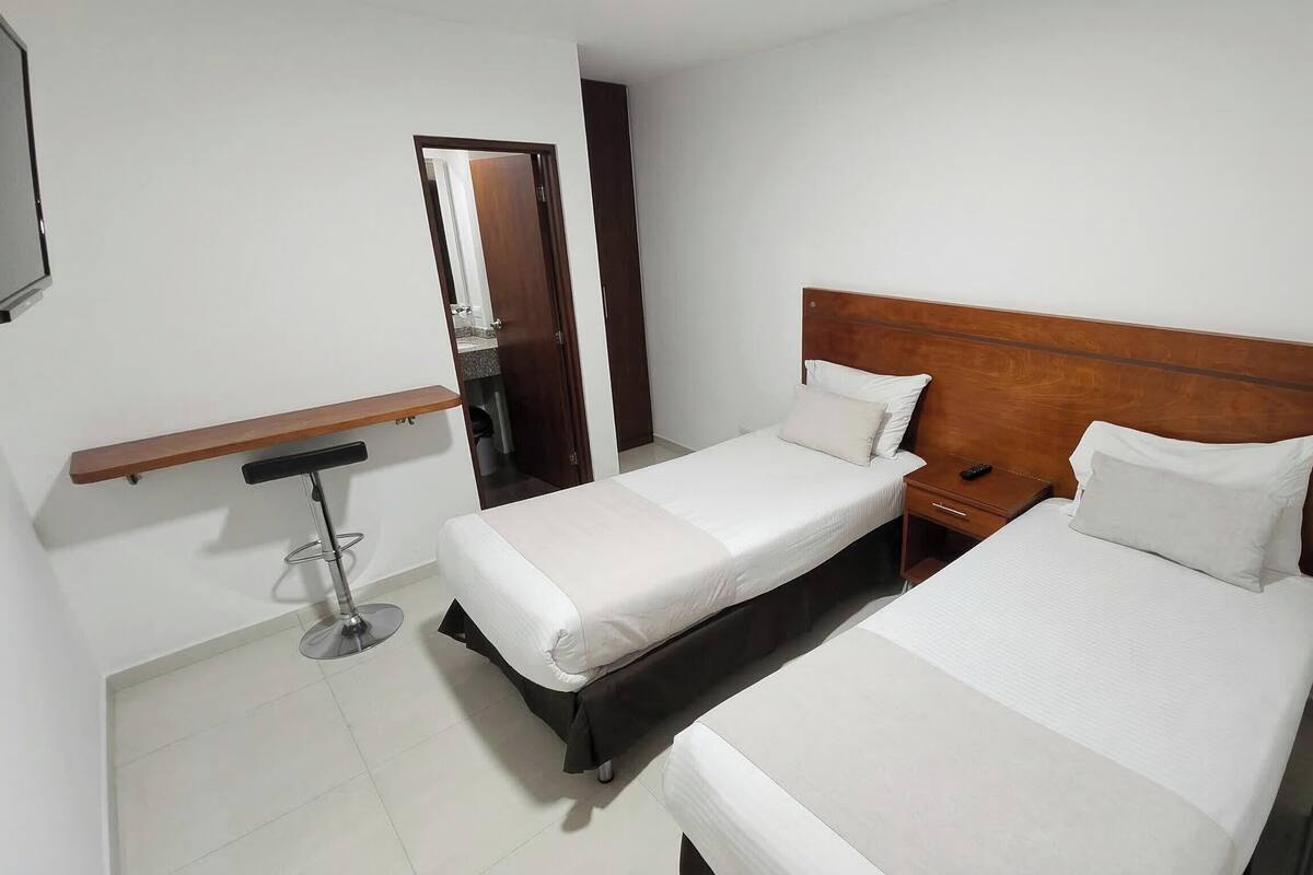 A hotel room with two beds, a wooden headboard, a bedside table with a remote control, and a small desk with a chair.