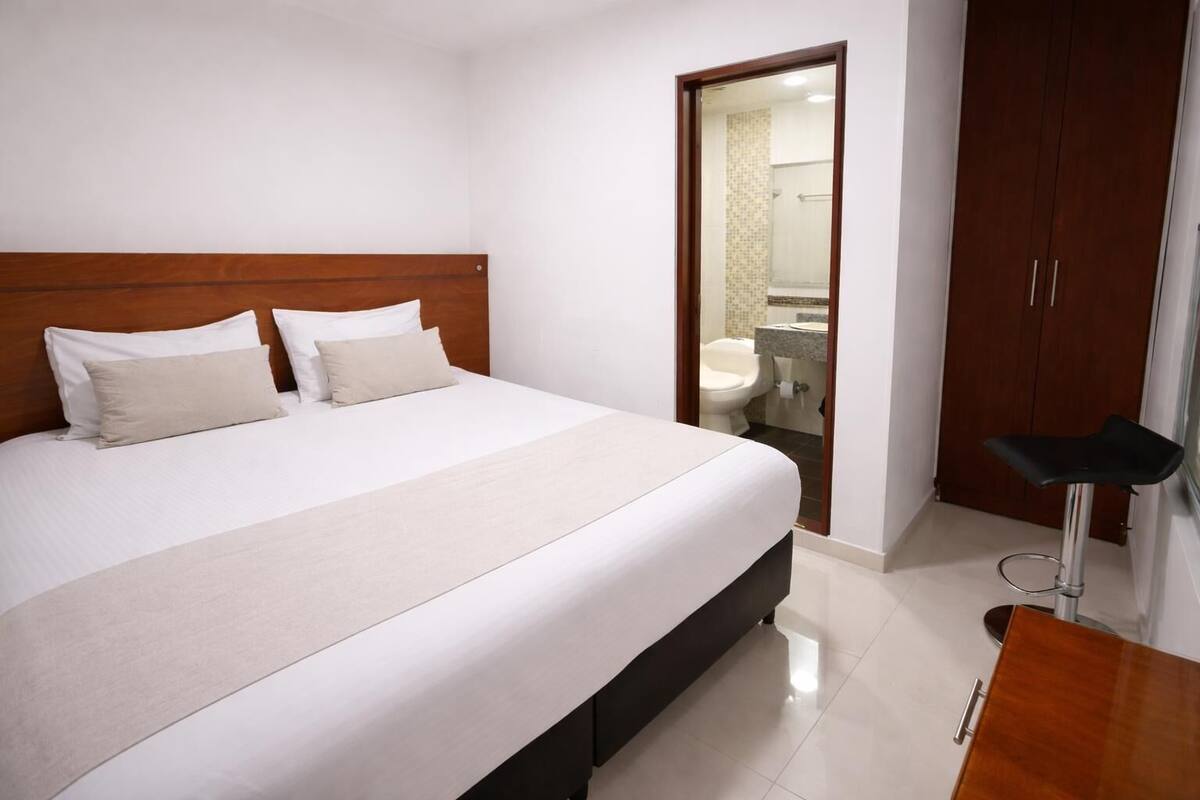 A hotel room with a bed, a wooden headboard, a bathroom visible through an open door, and a small stool.