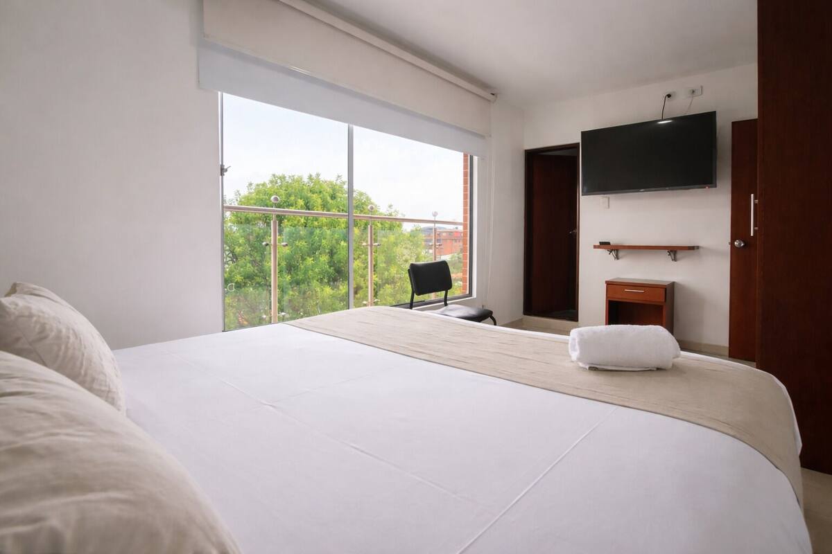 A hotel room with a large bed, a chair, a TV, and a balcony with a view.