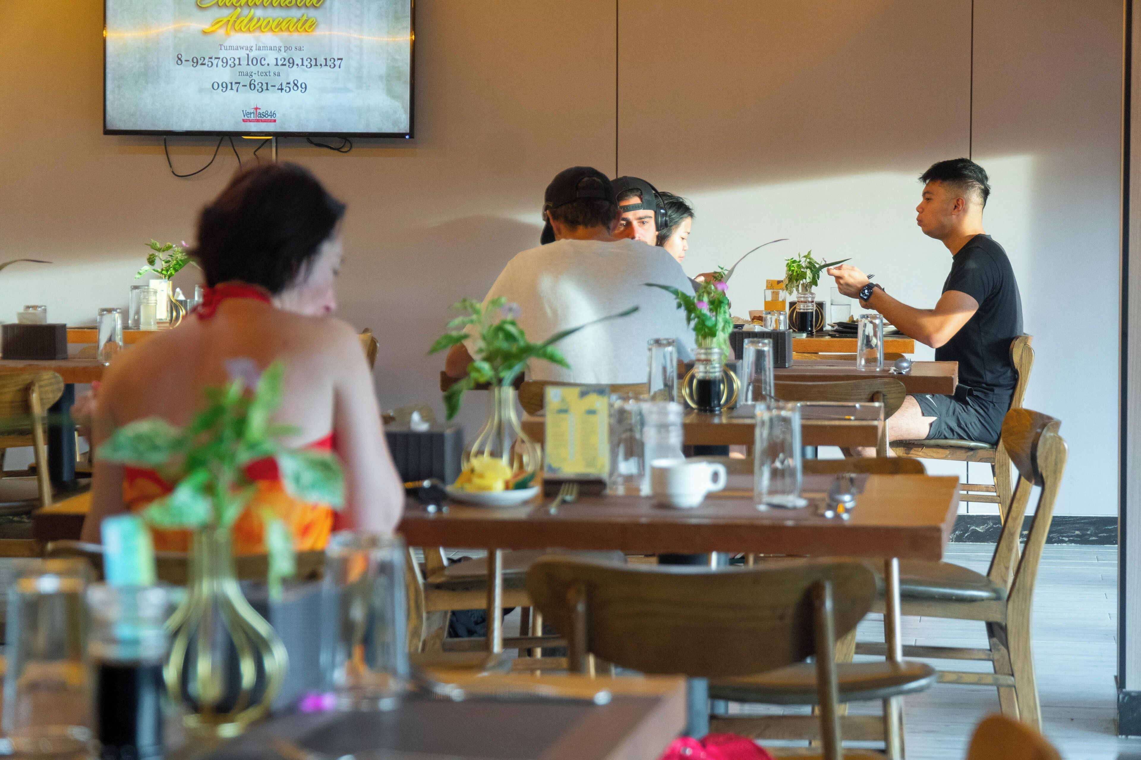 A restaurant with wooden tables and chairs, a TV screen displaying a menu, and various table settings including glasses and cutlery.