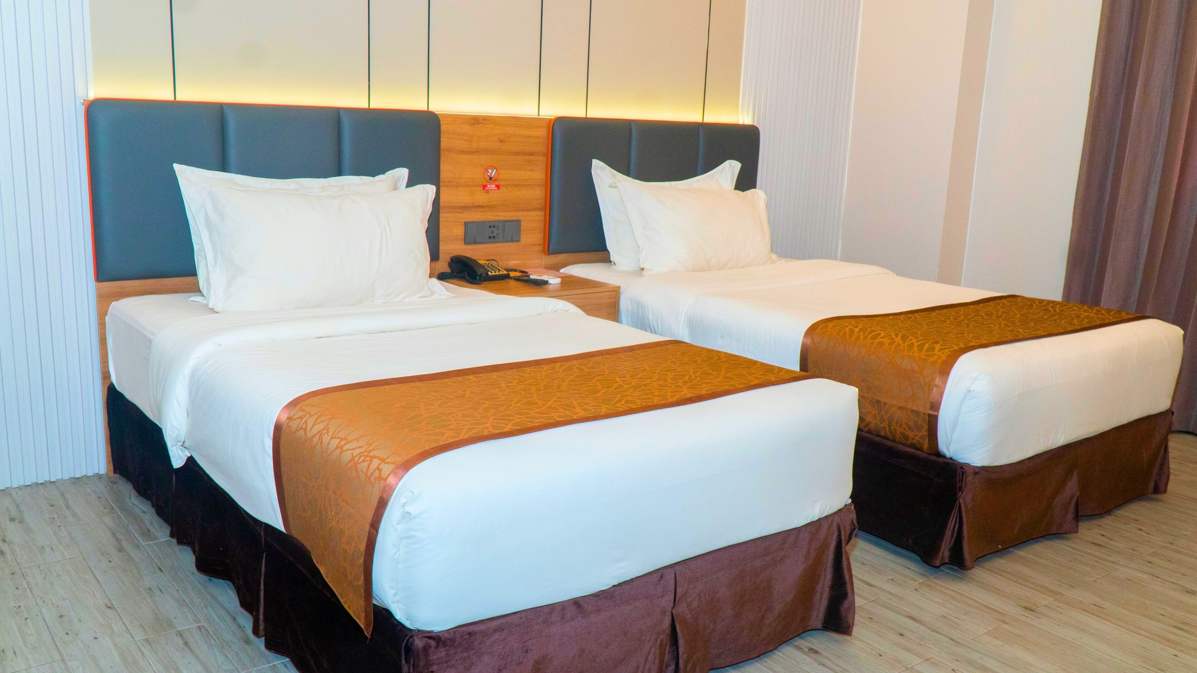 A hotel room with two beds, each with white linens and brown headboards. There is a wooden nightstand between the beds with a telephone and a remote control.