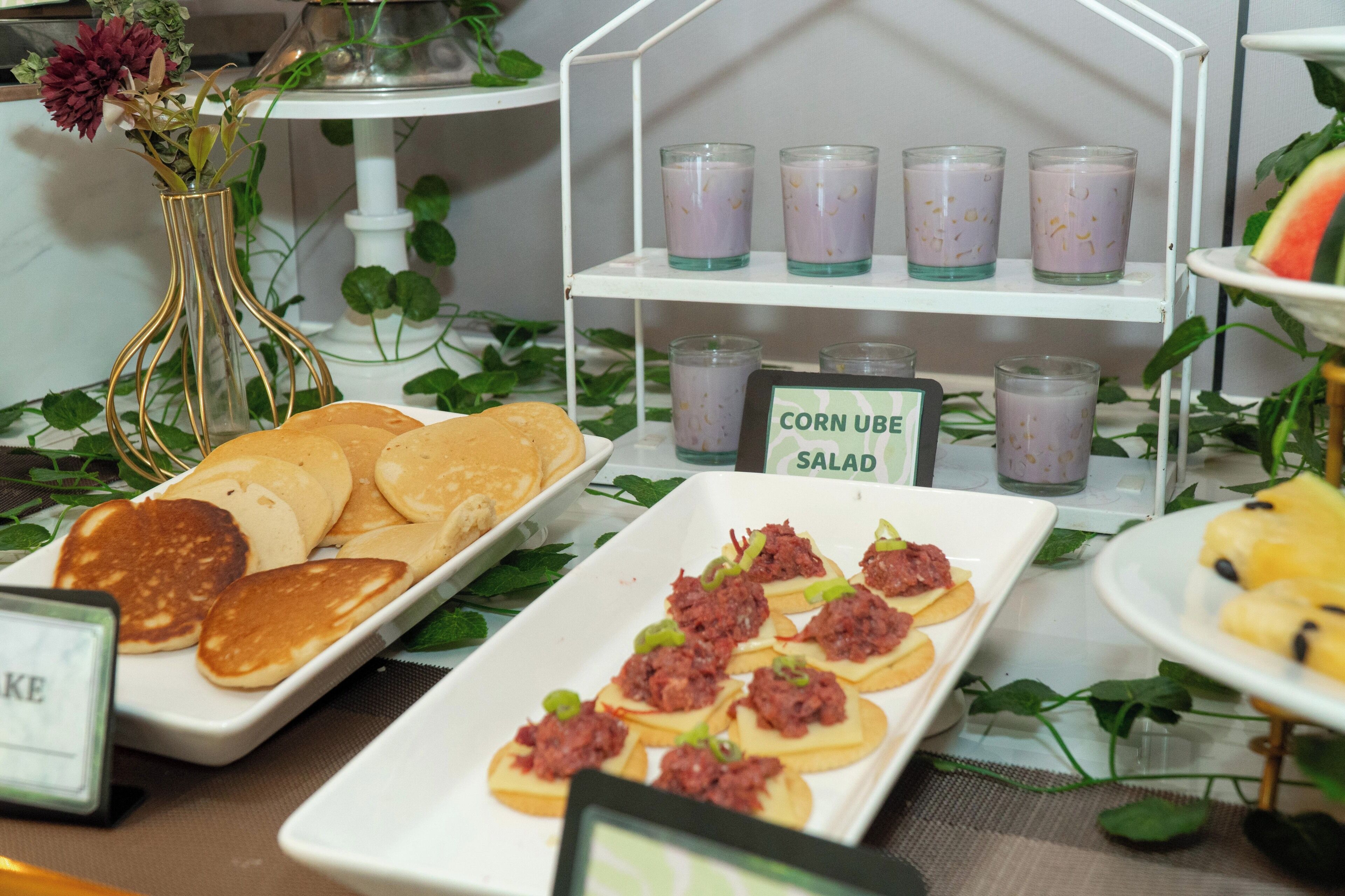 A buffet with various food items including pancakes, a salad, and a sign indicating 'CORN UBE SALAD'.