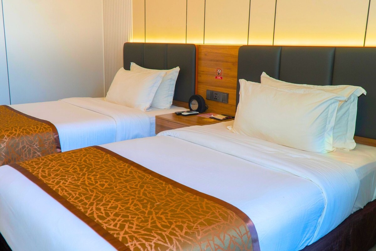 Une chambre d’hôtel avec deux lits, chacun agrémenté d’une couvre-lit à motifs et de coussins, une table de chevet avec un téléphone et une horloge, et un luminaire fixé au mur.