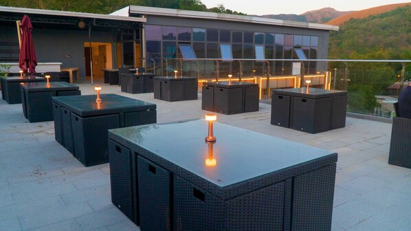 An outdoor seating area with tables and chairs, a glass railing, and a building in the background.