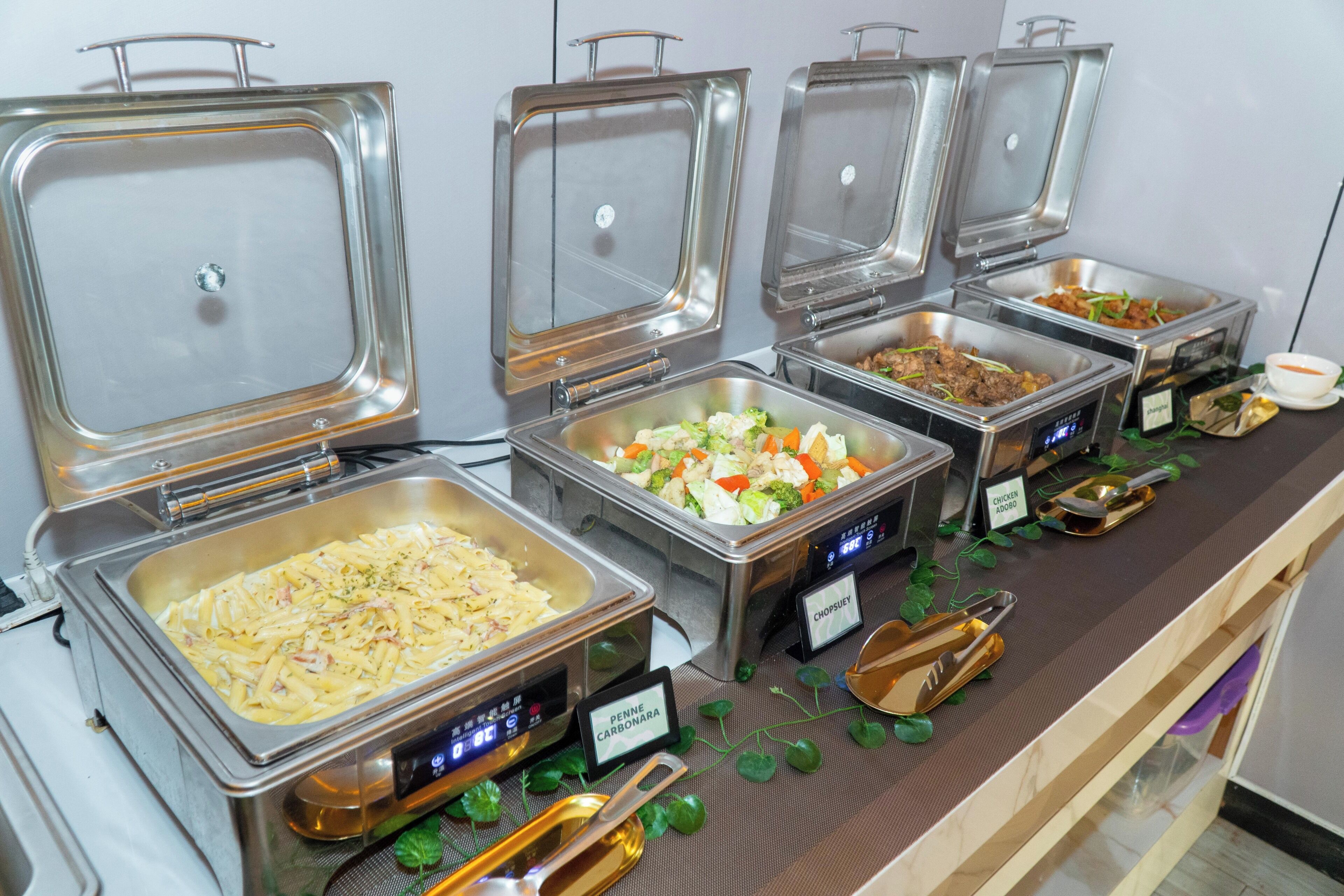 Un bufé con diversas opciones de comida, entre ellas pasta, ensaladas y carnes, identificadas con nombres como 