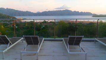 A rooftop terrace with sun loungers overlooking a coastal area with boats and a bridge.