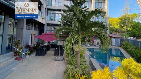 Exterior del Hotel Corona Viena con área de piscina, mobiliario exterior y una palmera.