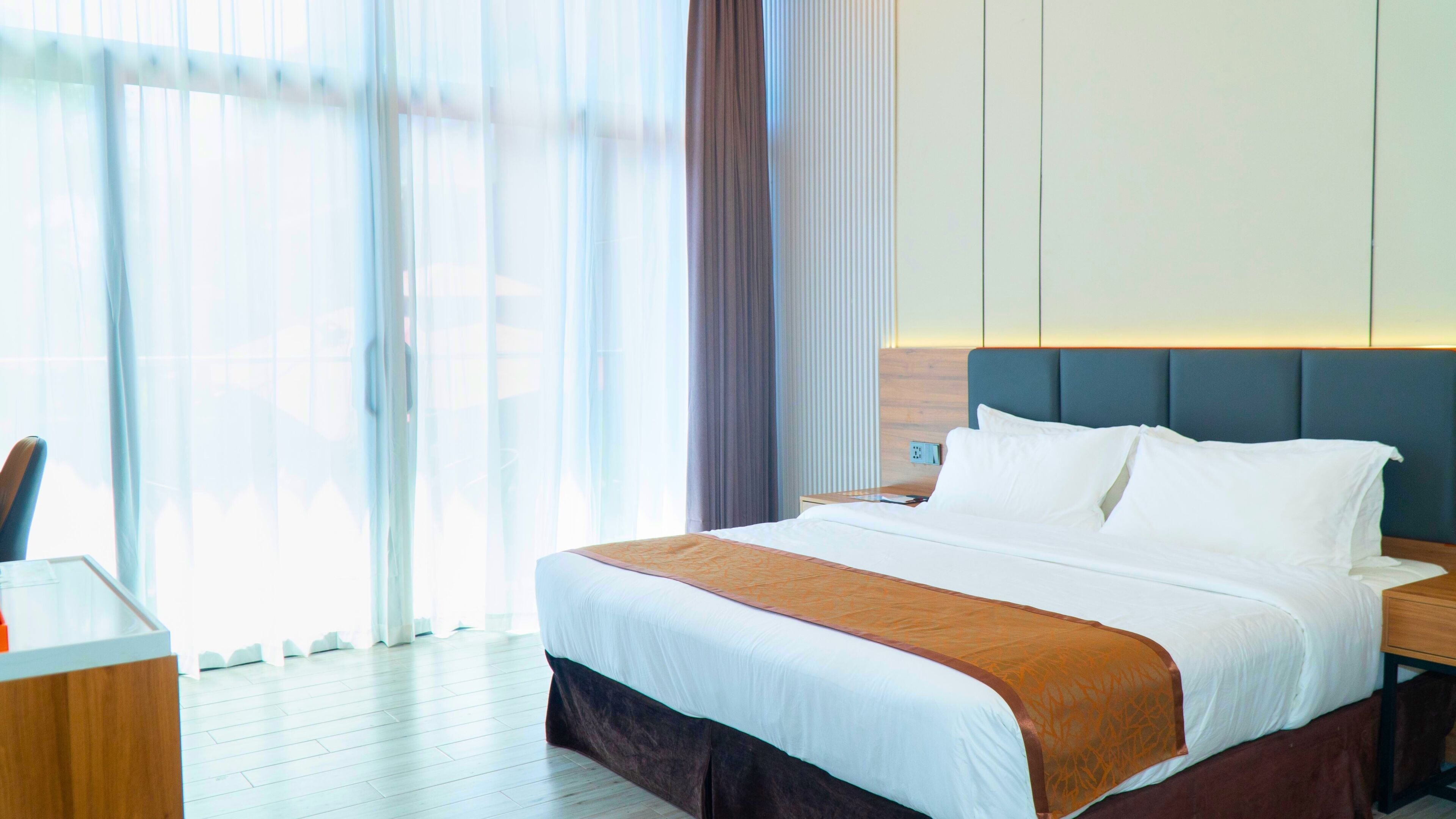 A hotel room with a large bed, a bedside table, a desk, and a window with sheer curtains.