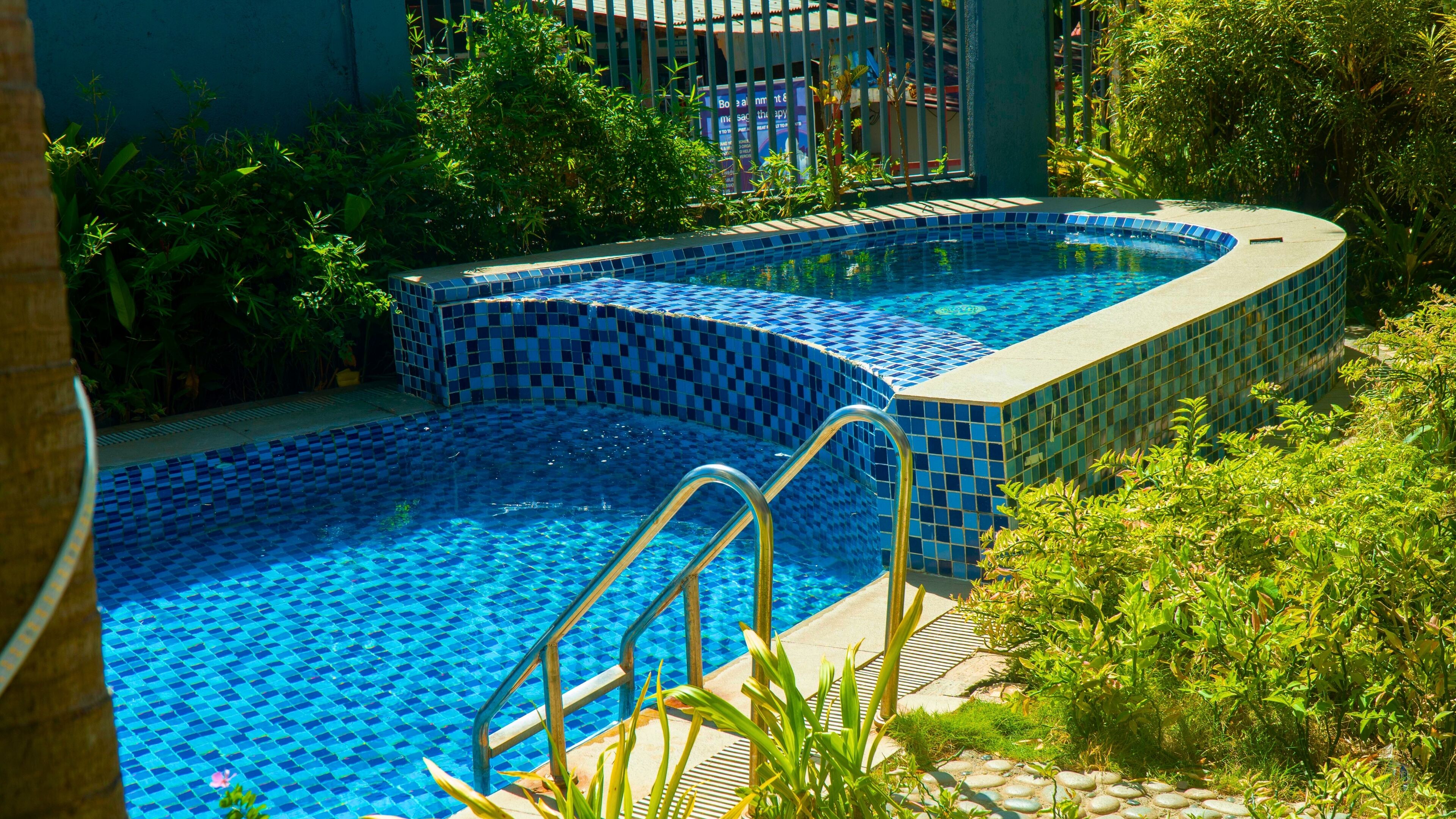 Ein Schwimmbecken mit blauen Mosaikfliesen, umgeben von Grünpflanzen und einem Metallzaun.
