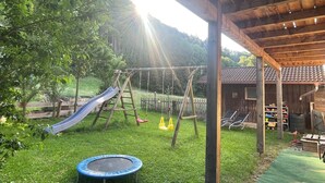 A wooden play structure with a slide and swings, a trampoline, and a covered area with a tiled roof. - Ferienwohnung Bergstation im Dopplerhof 2.OG 100qm, 2 Schlafzimmer (Piding)