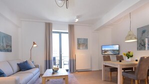 Ein modernes Wohnzimmer mit einer Couch, Couchtisch, Esstisch und Stühlen. An der Wand ist ein Fernseher, auf dem Tisch eine Pflanze und an den Wänden Kunstwerke.