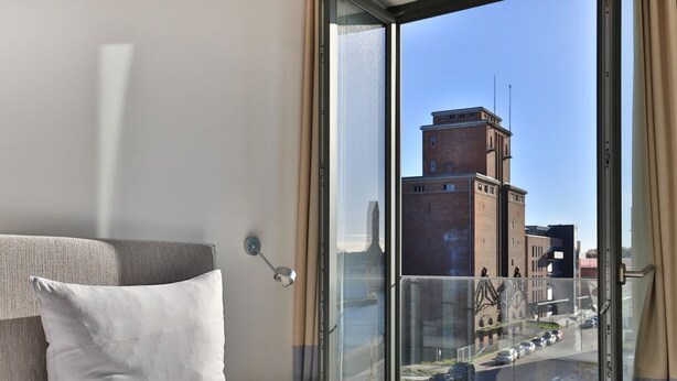Ein modernes Hotelzimmer mit großem Fenster, einem Bett mit weißer Bettwäsche und Blick auf eine Stadtlandschaft, darunter ein hohes Backsteingebäude.