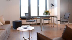 A modern living room with a sofa, a coffee table, and a dining table with chairs. There is a large window with blinds, a clock on the wall, and a lamp on the table.
