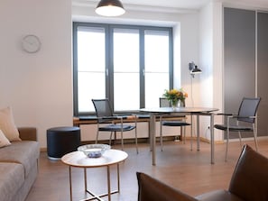 A modern living room with a sofa, a coffee table, and a dining table with chairs. There is a large window with blinds, a clock on the wall, and a lamp on the table.