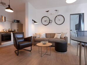 A modern living room with a sofa, a round coffee table, a black leather chair, and a dining table. A bicycle is mounted on the wall.