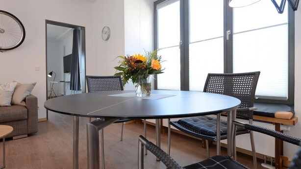 A modern living room with a round dining table, black woven chairs, a sofa, and a large window with blinds.