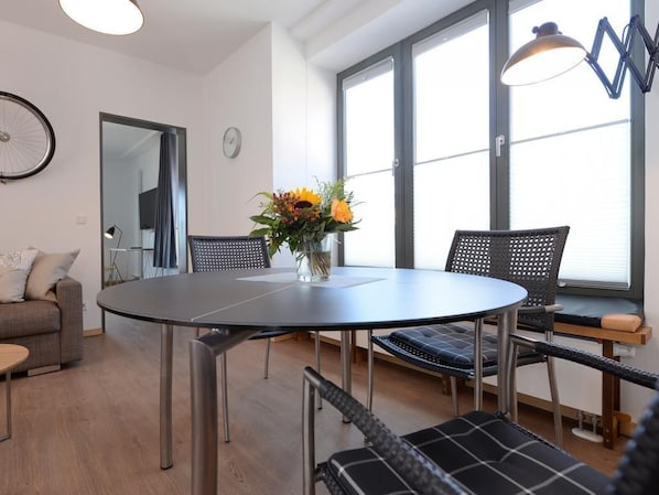 Ein modernes Wohnzimmer mit einem runden Esstisch, schwarzen gewobenen Stühlen, einer Couch und einem großen Fenster mit Jalousien.