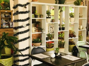 Una habitación con una estantería blanca llena de plantas en macetas, una biblioteca de madera con libros y adornos, y una mesa con menús.