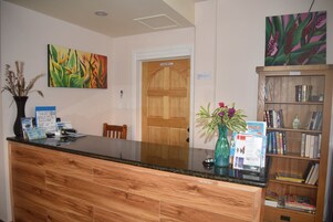 Un mostrador de recepción con encimera de madera, un jarrón con plantas secas, un cuadro y una estantería con libros.