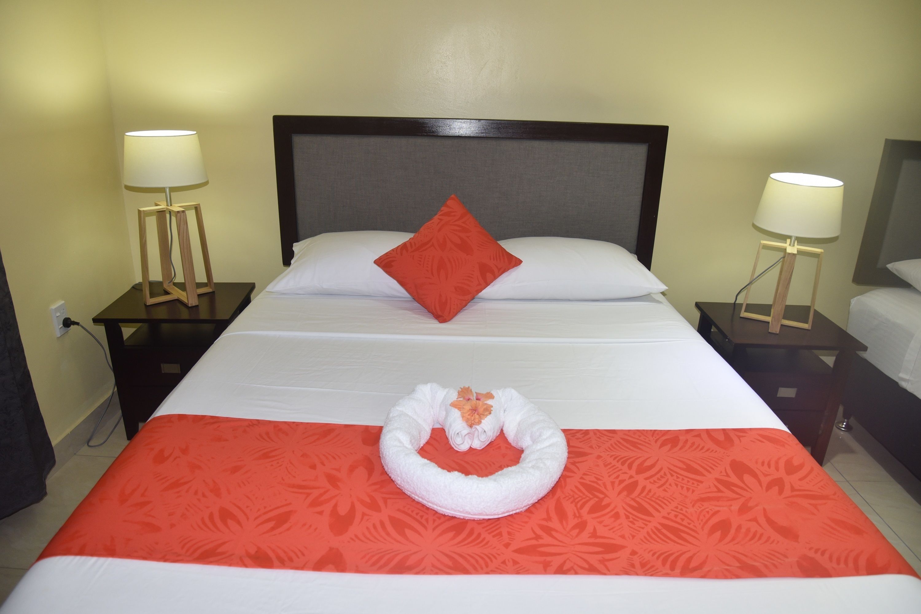 Una cama bien hecha con un almohadón rojo, una toalla blanca y una colcha de rayas rojas y blancas. Hay dos mesitas de noche con lámparas.