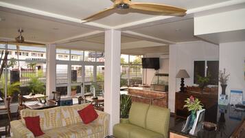 A spacious living area with a sofa, armchair, and a coffee table. There is a ceiling fan, a TV, and a window with a view of buildings and greenery outside.