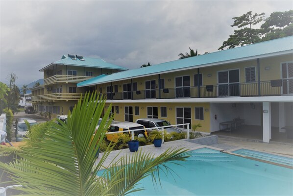 Un hotel con varios balcones, piscina y coches aparcados.