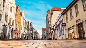 Una strada acciottolata fiancheggiata da edifici storici, alcuni con vetrine, sotto un cielo azzurro limpido.